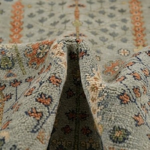May include: A light blue area rug featuring a repeating pattern of floral motifs in shades of orange, dark blue, and cream. The rug has a dense, tufted pile texture and is folded in the centre, revealing the intricate woven design and soft fibres.