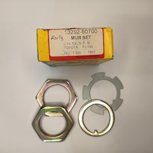 Op de afbeelding: Een Toyota auto-onderdelenset in een geel-witte doos met het label 13202-90700 MUR SET. De set bevat twee zeshoekige metalen moeren met een metallic afwerking, één ronde metalen ring en één grijze metalen borgring met vier lipjes.