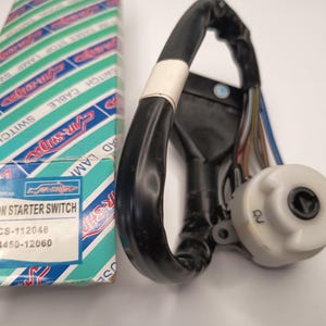 Op de afbeelding: Een ontstekingsstartschakelaar met een witte cilindrische behuizing en een zwarte kabelboom in zwarte beschermende buizen. De schakelaar ligt naast de originele verpakking, die blauwgroen en wit is met de tekst IGNITION STARTER SWITCH, CS-112048 en 84450-12060 op het etiket.