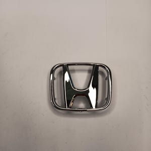 May include: A chrome-finished Honda emblem featuring the iconic H logo enclosed within a rounded rectangular frame. The badge has a reflective, metallic surface and includes small mounting pins on the back side for secure vehicle attachment.
