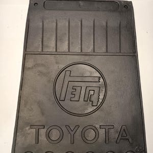 Op de afbeelding: Een zwarte rechthoekige rubberen spatlap met het Toyota-logo in reliëf in een cirkel in het midden. Het woord TOYOTA is in hoofdletters onder het logo gegoten. Het bovenste gedeelte heeft verticale ribbels en twee montagegaten.