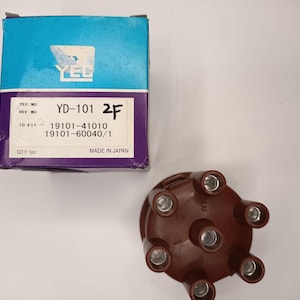 May include: A brown distributor cap with six metal terminals and a corresponding blue and purple cardboard box. The box features the YEC logo, the text YD-101, 2F, and the reference numbers 19101-41010 and 19101-60040/1, with Made in Japan printed.