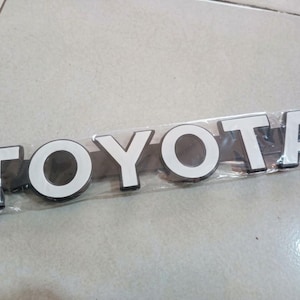 Può includere: Un emblema per veicoli Toyota caratterizzato da lettere in stile blocco con finitura bianca e bordo scuro. L'emblema è racchiuso in una custodia protettiva in plastica trasparente, che mostra chiaramente il marchio in orizzontale.