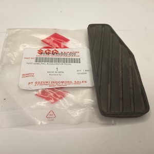 May include: A black rubber accelerator pedal pad with vertical grooves, positioned next to a clear plastic packaging bag featuring the Suzuki logo, the text 49481M60B00-000, PART NAME PAD, ACCELERATOR PEDAL, and PT SUZUKI INDOMOBIL SALES.
