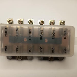 以下が含まれることがあります： 半透明の白いプラスチックカバーが付いた長方形の自動車用ヒューズブロック。カバーには、TURN 15A、WIPER 15A、METER 5A、SPARE 20A、HORN 20A、LIGHT 20Aの黒いラベルが印刷されています。上下の端に金属製のネジ端子が見えます。