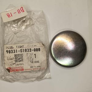 May include: A circular metal engine plug with a smooth, reflective finish, positioned next to a clear plastic bag containing a label that reads PLUG, TIGHT 90331-51032-000 and includes the Daihatsu Engine Parts logo and a QR code.