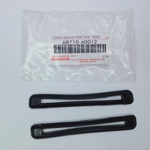 Op de afbeelding: Twee zwarte, rechthoekige, holle kunststof onderdelen voor een voertuigdeurscharnierconstructie. Ze zijn verpakt in een doorzichtige plastic zak met een wit label met de tekst: CUSHION, HINGE ASSY FRONT DOOR - FEMALE, 68710-60012, TOYOTA GENUINE PARTS.