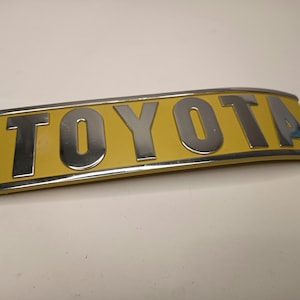 Op de afbeelding: Een gebogen, rechthoekig Toyota-embleem met verhoogde, gepolijste chromen letters tegen een gladde, mosterdgele achtergrond. Het stuk is omlijst door een dunne, metalen chromen rand die de zachte boog van het ontwerp volgt.