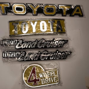 Op de afbeelding: Een verzameling van vijf auto-emblemen in plastic verpakking. De set bevat twee TOYOTA-badges, twee TOYOTA Land Cruiser-badges met zilveren letters op een zwarte achtergrond en één 4 WHEEL DRIVE-badge met een rood cijfer vier op een gouden achtergrond.