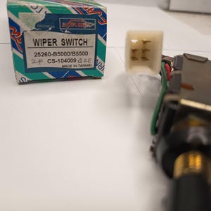 以下が含まれることがあります： 黒いハンドル、金属製の本体、6本の金属ピンが付いた白いプラスチック製コネクタを備えたワイパースイッチ。横には、WIPER SWITCH、25260-B5000/B5500、CS-104009 G2E、MADE IN TAIWANと書かれた青、白、ターコイズブルーの箱があります。