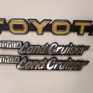 以下が含まれることがあります： 3つの自動車用エンブレムのセット。一番上のエンブレムは、黒い縁取りの黄色いブロック体でTOYOTAと書かれており、黒いテクスチャの背景に配置されています。その下には、TOYOTA Land Cruiserと書かれた同一の銀色の金属製エンブレムが2つ並んでいます。