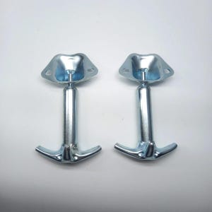 Op de afbeelding: Twee zilverkleurige metalen hardware-onderdelen, elk voorzien van een platte, driehoekige montageplaat met twee ronde schroefgaten aan de bovenkant, verbonden met een verticale cilindrische paal die eindigt in een gebogen dubbele haakvoet.