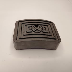 Op de afbeelding: Een rechthoekige zwarte rubberen pedaalafdekking met een verhoogd geometrisch patroon op het oppervlak. Het ontwerp bestaat uit een centraal cirkelvormig element omringd door concentrische rechthoekige randen voor een goede grip.