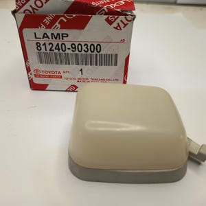 Op de afbeelding: Een beige rechthoekige autolamp met een grijze voet en een klein aansluitlipje, geplaatst voor een wit en rood Toyota originele onderdelen doos met de tekst LAMP 81240-90300 en Toyota Motor Thailand Co. Ltd. Made in Thailand.