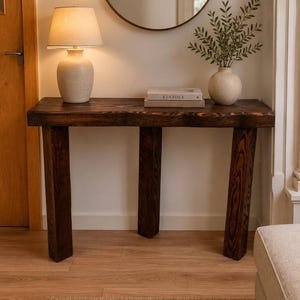 May include: A dark brown wooden console table with four thick legs, topped with a beige ceramic table lamp, a stack of two books with the word KINFOLK visible on the spine, and a white ceramic vase containing green leafy branches.