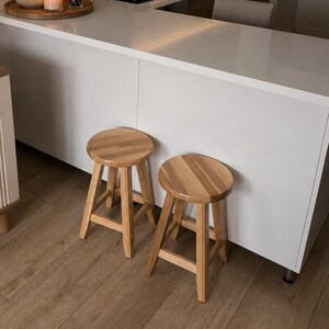 Peut inclure: Un intérieur de cuisine avec un îlot à comptoir blanc et deux tabourets ronds en bois. La cuisine comprend des armoires blanches, une plaque de cuisson noire et un évier. Des éléments décoratifs incluent un plateau en bois avec un vase.