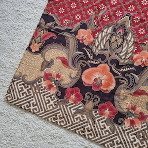Op de afbeelding: Een textielstof met een donkerbruine achtergrond met grote oranje en rode orchideeën, wervelende bladpatronen en een geometrische rand in crème en zwart. Het bovenste gedeelte toont een rood veld met kleine witte bloemmotieven.