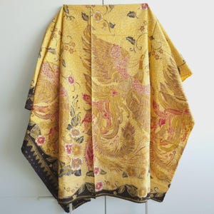 Op de afbeelding: Een gele stof met een gedetailleerde batikprint van feniksen en bloemmotieven in bruine, rode en oranje tinten. Het textiel vertoont overal ingewikkelde lijnen en patronen, met een donkerbruine rand langs de onderkant.