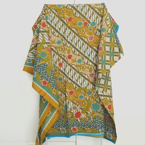 Puede incluir: Un tejido rectangular con fondo amarillo mostaza que presenta un patrón batik repetitivo de líneas onduladas diagonales, motivos florales en rojo y azul, y secciones de cuadrícula geométrica. La tela tiene un borde azul brillante.