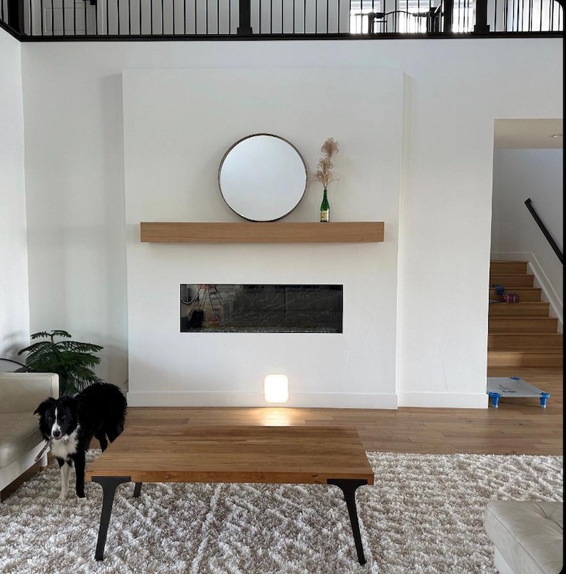 May include: A modern living room with a white fireplace, a round mirror, a wooden coffee table, and a black and white dog. The fireplace has a black hearth and a wooden mantel.