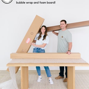 May include: Two people are packing a large wooden table for shipping. The table is wrapped in bubble wrap and secured with cardboard boxes. The text on the box reads "FRAGILE".