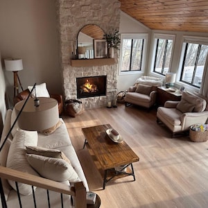 May include: A living room with a stone fireplace, a white couch, two armchairs, a coffee table, and a wooden ceiling. The fireplace has a fire burning in it and there are windows on the right side of the room.