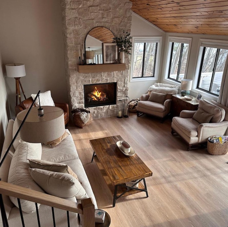 May include: A living room with a stone fireplace, a white couch, two armchairs, a coffee table, and a wooden ceiling. The fireplace has a fire burning in it and there are windows on the right side of the room.