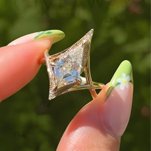 Puede incluir: Un anillo de oro con una piedra central en forma de cometa con superficie facetada. El engaste incluye un marco triangular texturizado en un lado de la piedra, acentuado con pequeñas piedras claras y brillantes a lo largo de los bordes.
