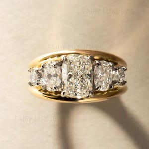May include: A gold ring featuring a central cushion-cut diamond flanked by two oval-cut diamonds and two smaller round-cut diamonds, all set in a polished yellow gold band. The text FAIRY LUX JEWEL is visible in the background.