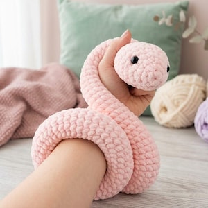 May include: A soft, light pink crochet snake toy made from chunky, textured yarn. The plush snake features a rounded head with two small, smooth black plastic eyes and a long, coiled body that wraps around the wrist.