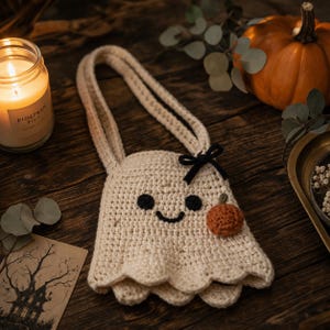 May include: A cream-colored crochet bag shaped like a ghost, featuring two long handles, black yarn eyes, a black yarn smile, a small black ribbon bow, and a tiny orange crochet pumpkin accent attached to the front of the bag.