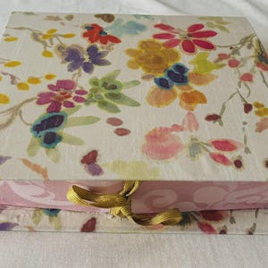 Könnte beinhalten: Eine quadratische Schachtel mit Klappdeckel, die ein Aquarell-Blumenmuster in Rosa-, Gelb-, Lila- und Rottönen auf weißem Grund aufweist. Der Boden der Schachtel ist hellrosa mit einem dezenten Ton-in-Ton-Muster und einem goldenen Bandverschluss.