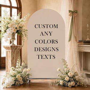 May include: A tall, arched, off-white display sign featuring the black text CUSTOM ANY COLORS DESIGNS TEXTS. The sign is positioned upright on a thin metal base, serving as a blank canvas for event signage, surrounded by white floral arrangements.