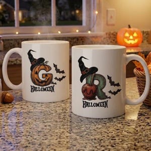 May include: Two white ceramic mugs featuring Halloween-themed graphics. The left mug displays a pumpkin shaped like the letter G wearing a witch hat, with the word HALLOWEEN below. The right mug shows a candy apple shaped like the letter R.