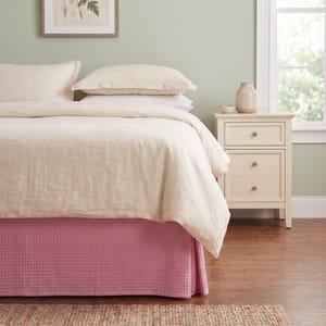 May include: A bed featuring a textured pink bed skirt with a waffle pattern, topped with a cream-colored duvet and two matching pillows. Beside the bed stands a cream-colored wooden nightstand with three drawers and small round knobs.