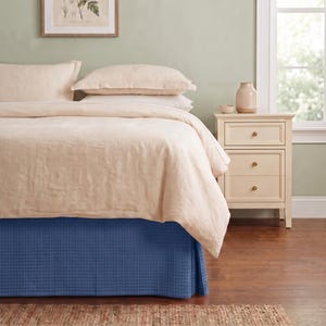 May include: A bed featuring a light beige duvet cover and matching pillows, paired with a blue waffle-weave bed skirt. Beside the bed sits a light-colored wooden nightstand with three drawers, each equipped with a small, round, metallic knob.