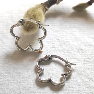 May include: A pair of silver hoop earrings shaped like five-petalled flowers. Each earring features a smooth, polished metallic finish and a hinged closure mechanism, creating a continuous outline of a blossom in a simple, open-frame design.