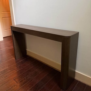 Pode incluir: Uma mesa console de madeira castanho-escura com uma superfície superior longa e retangular e dois painéis laterais largos e sólidos. A mesa tem um acabamento liso e mate com cantos arredondados, criando uma estrutura limpa e unificada.