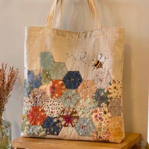 May include: A beige canvas tote bag featuring a patchwork design of various floral and patterned fabric hexagons. The lower portion is covered in these quilted shapes, while an embroidered bumblebee with a dotted flight path sits above.