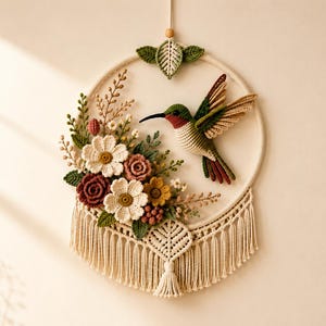 May include: A circular wall hanging featuring a crocheted hummingbird in flight, surrounded by a cluster of crocheted flowers and foliage in shades of red, white, and green. The piece is mounted on a wooden hoop with cream-colored macrame fringe.