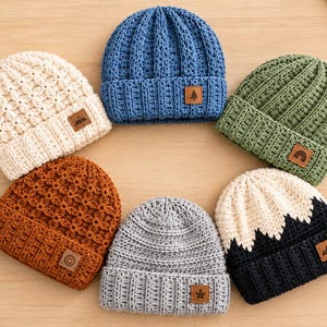 May include: Six crocheted beanies arranged in a circle, featuring various colors including cream, blue, green, rust, gray, and a black and white mountain pattern. Each hat has a folded brim and a small square brown leather-like patch with an icon.