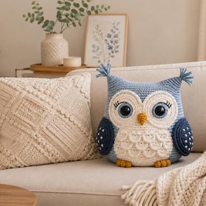 Pode incluir: Uma almofada de crochê em forma de coruja com corpo azul claro, rosto branco, olhos pretos grandes, um pequeno bico amarelo e asas azul-escuras com pontos brancos. Ao lado, há uma almofada quadrada creme com um padrão geométrico de tricô.