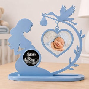 Può includere: Una cornice decorativa in legno azzurro con la sagoma di una persona incinta e un ritaglio circolare per un'ecografia. Una cicogna con un fagotto pende sopra una cornice a forma di cuore contenente la foto di un bambino sorridente.