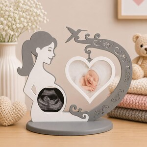 Può includere: Un espositore in legno grigio e bianco con la sagoma di una persona incinta, un'ecografia circolare nella zona della pancia e una cornice a forma di cuore appesa a un ramo curvo con un colibrì, contenente la fotografia di un neonato.