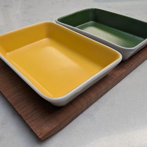 Może przedstawiać: Dwa prostokątne ceramiczne naczynia do pieczenia, jedno żółte i jedno zielone, leżą na drewnianej desce do serwowania. Naczynia mają białe obramowania i są puste. Deska jest ciemnobrązowa.