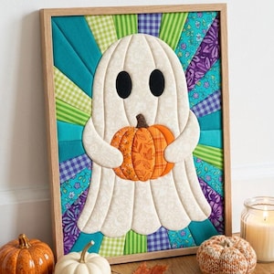 May include: A framed fabric wall art piece featuring a quilted white ghost with black oval eyes holding an orange pumpkin. The background consists of radiating fabric strips in shades of teal, green, purple, and blue with various patterns.