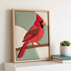 May include: A framed fabric wall art piece featuring a red cardinal bird perched on a brown branch. The bird has a black face mask and an orange beak. The background consists of geometric shapes in shades of sage green and cream fabric.