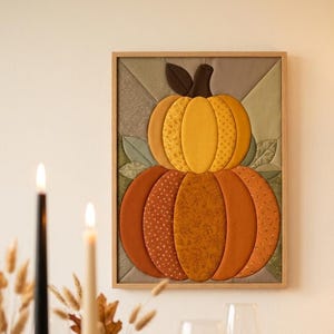 May include: A rectangular fabric wall art piece featuring a stacked pumpkin design in shades of yellow and orange, framed in light wood. The pumpkins are constructed from quilted fabric segments with subtle patterns, accented by green fabric leaves.