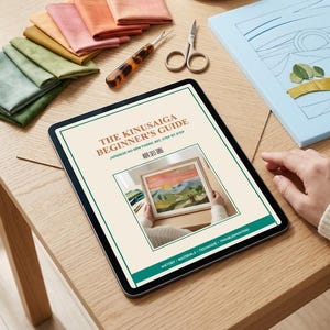 May include: A tablet displaying a digital guide titled The Kinusaiga Beginner's Guide, surrounded by fabric scraps in green and orange hues, a seam ripper, small scissors, a tea cup, and a light blue foam board with a partially completed fabric art design.