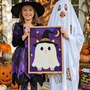 May include: A rectangular framed wall art piece featuring a fabric applique of a white ghost cat wearing a black witch hat. The background is a deep purple with layered wave patterns, accented by small yellow stars and a crescent moon in the corner.